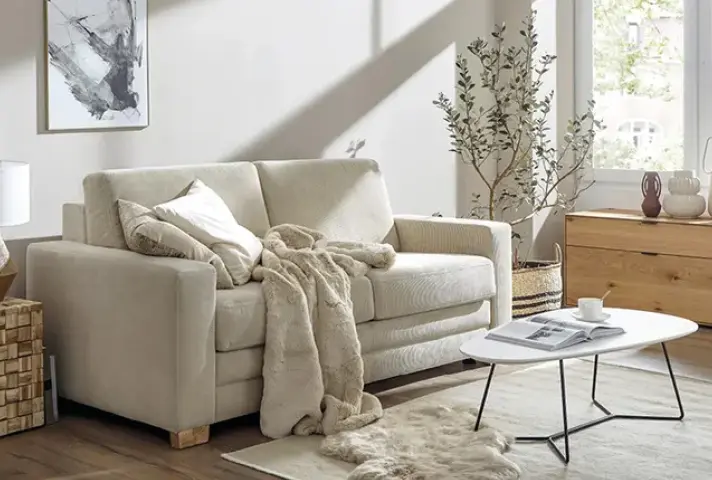 Beiges Zweisitzer Sofa mit Kissen und Decke, davor weisser ovaler Couchtisch mit Tasse und Zeitschrift auf Teppich, rechts Holzkommode und Pflanze am Fenster.